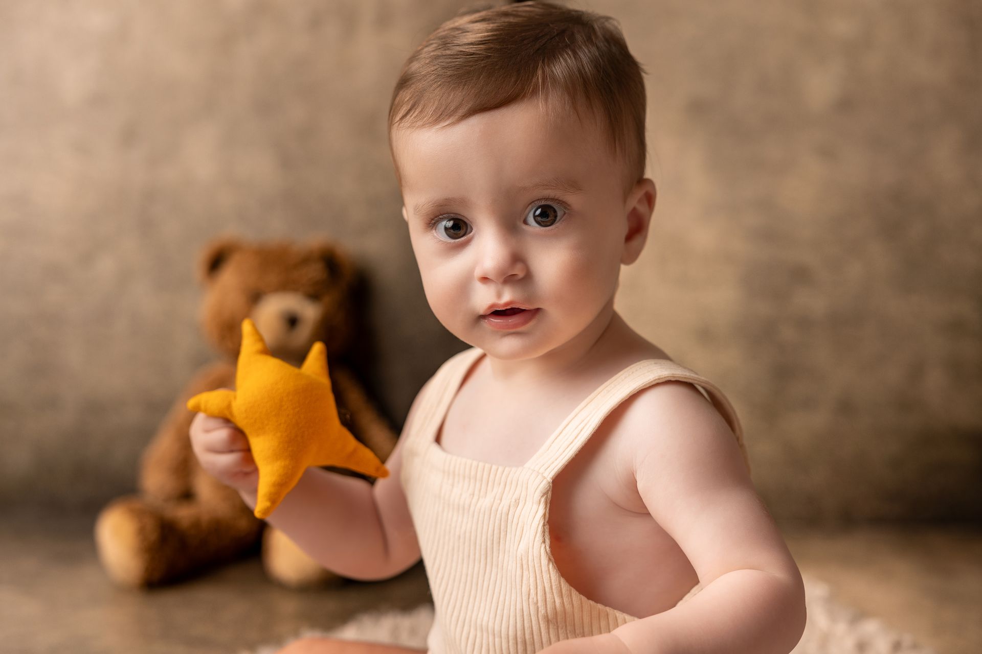 Bambino seduto in canottiera panna che tiene una stellina gialla, l'orsacchiotto accanto