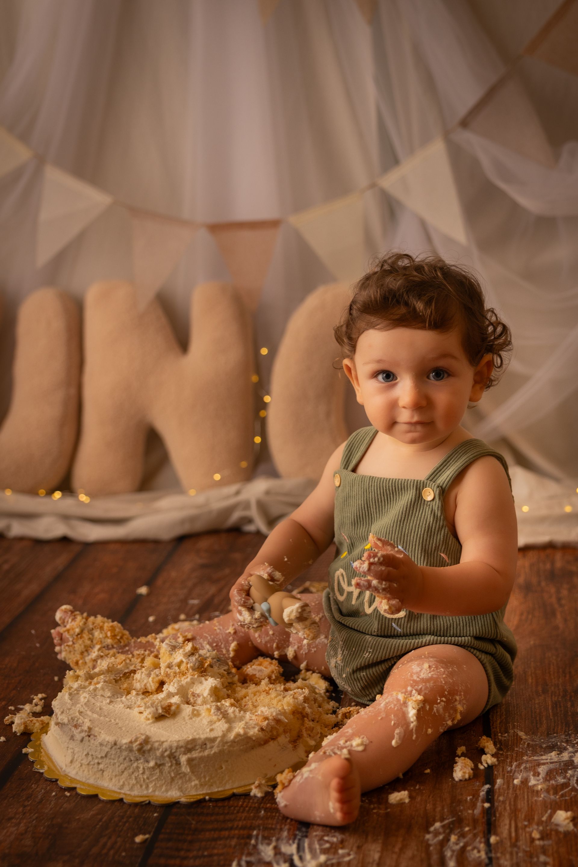 Bambino in tutina verde seduto accanto alla torta sbriciolata, lettere UNO e lucine sullo sfondo