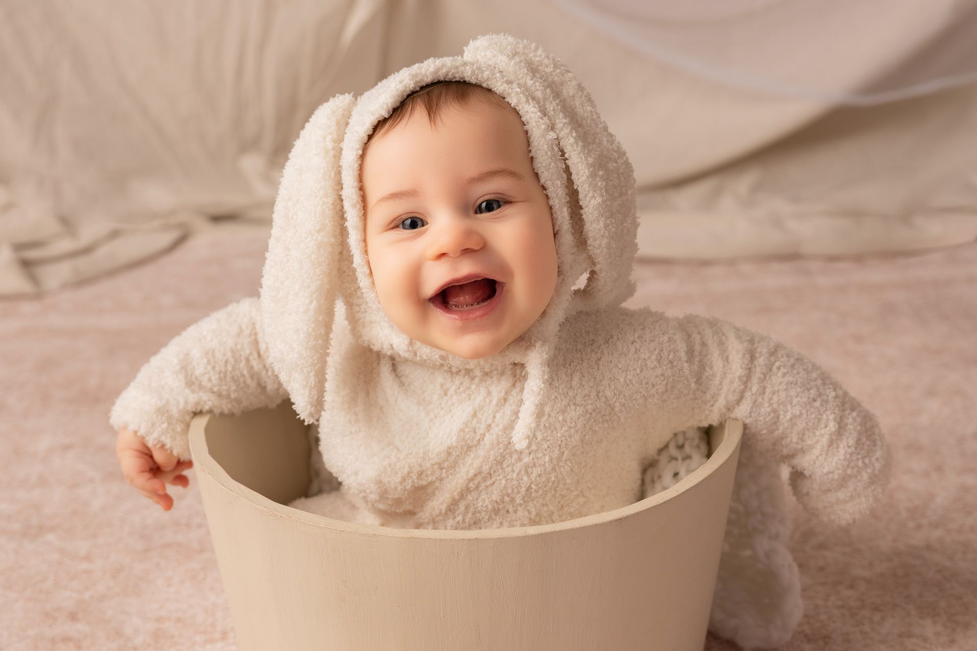 Bambino con tutina e cappuccio da coniglietto avorio ride dentro un mastello beige su fondo rosa