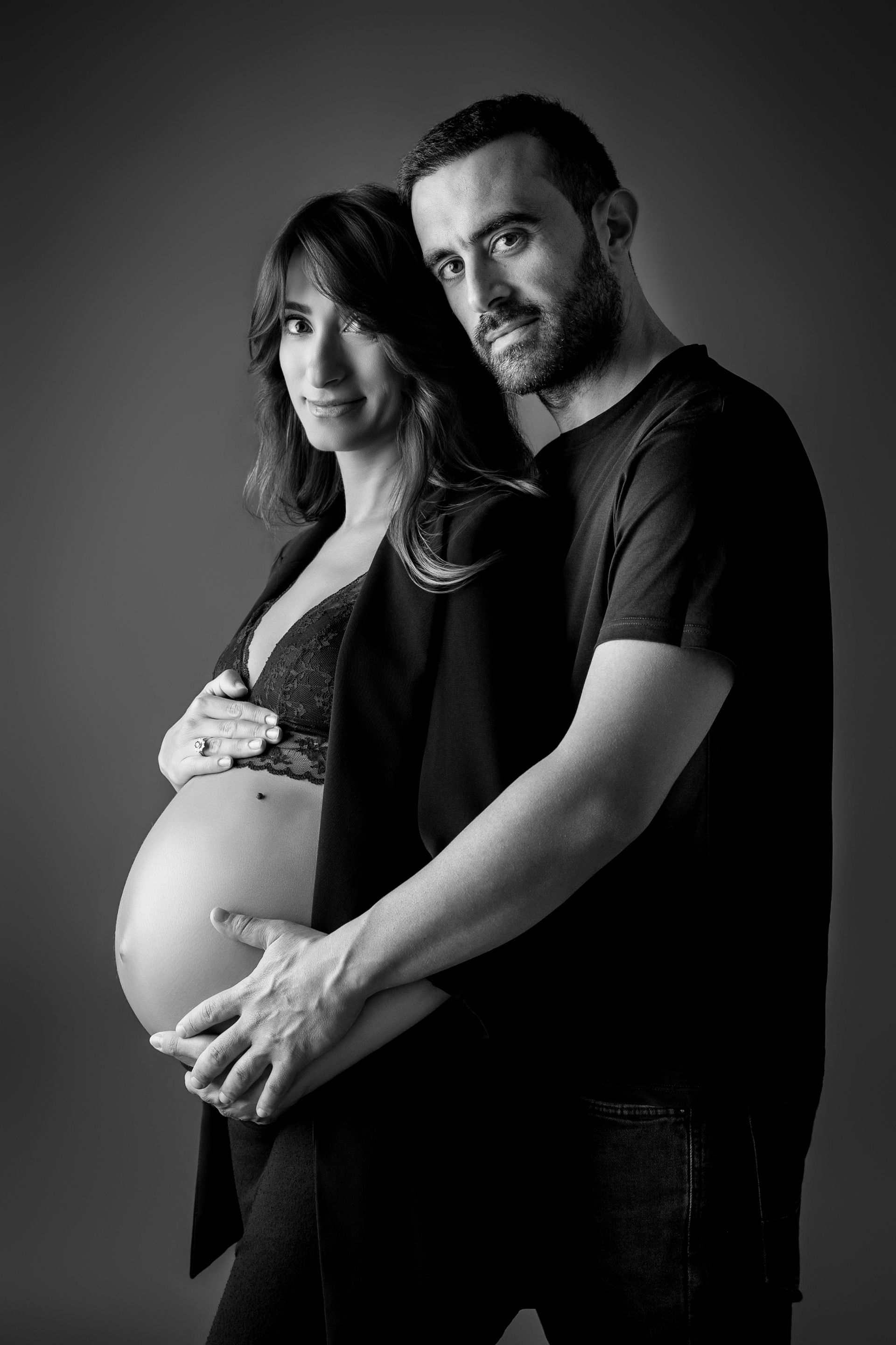 Compagno abbraccia da dietro la futura mamma in blazer nero, mani intrecciate sul ventre nudo in bianco e nero