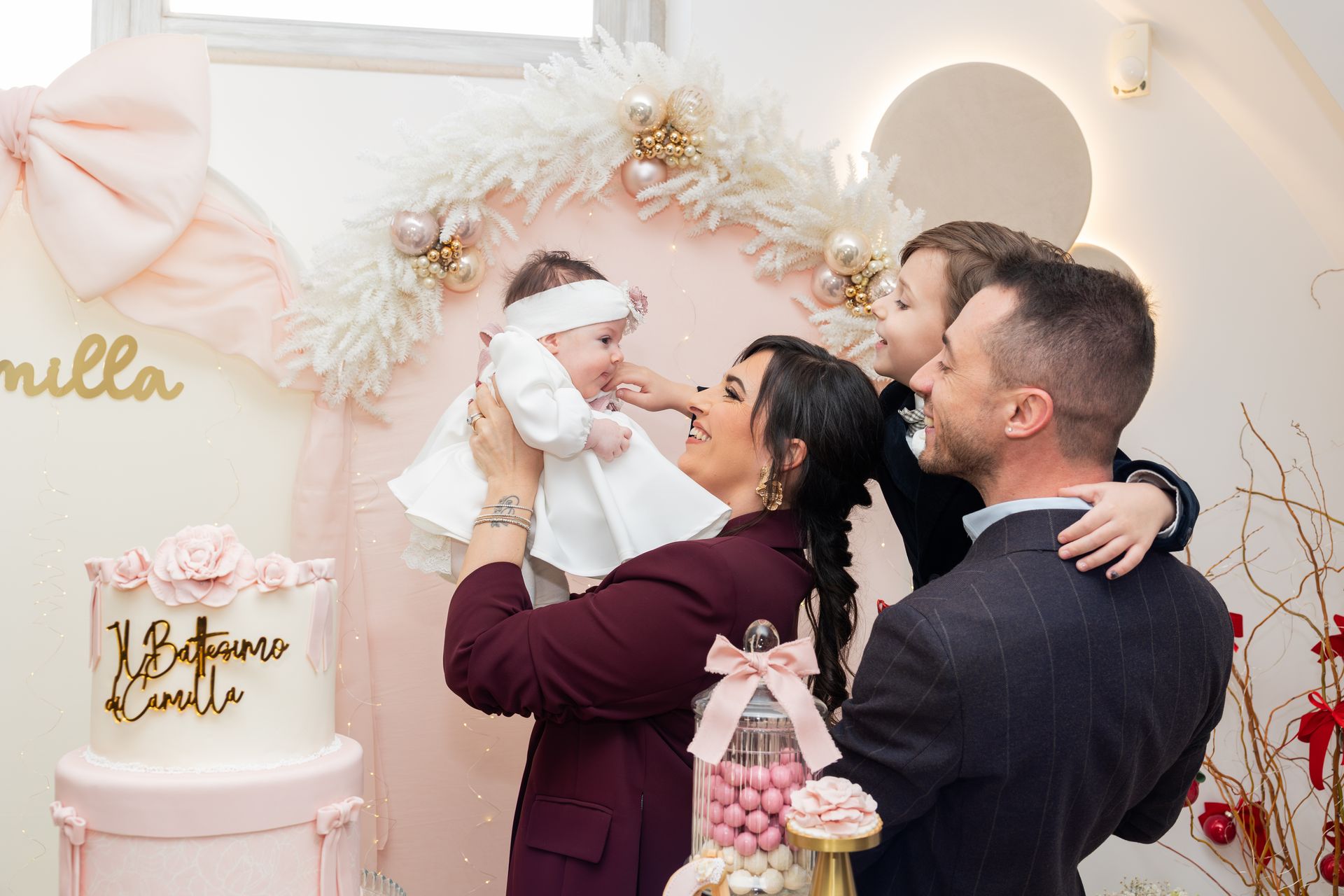 Mamma e papà sollevano la neonata in abito bianco davanti all'allestimento rosa del battesimo, con la torta e i fiori