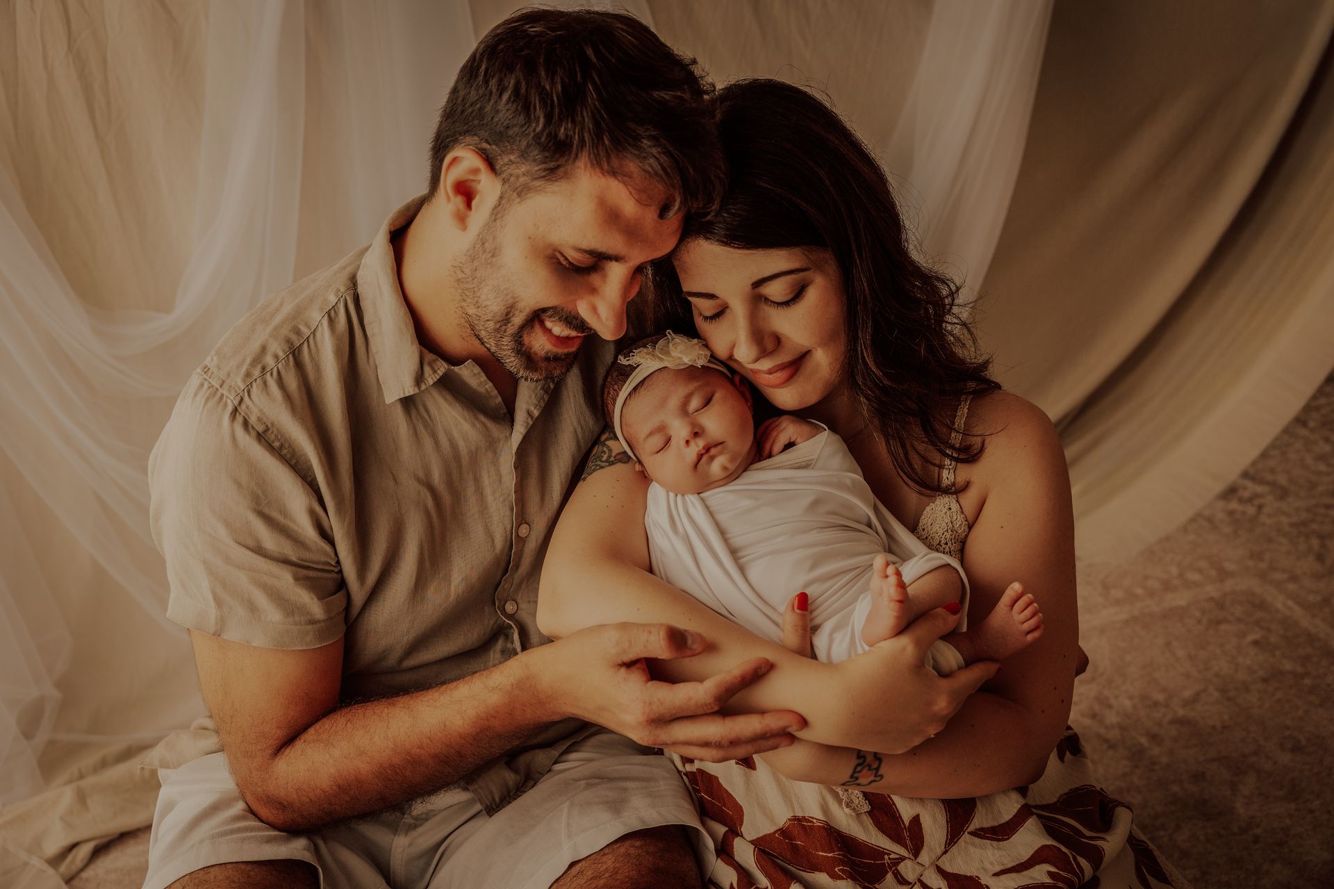 Mamma e papà seduti insieme sorreggono il neonato fasciato davanti a tende beige in studio