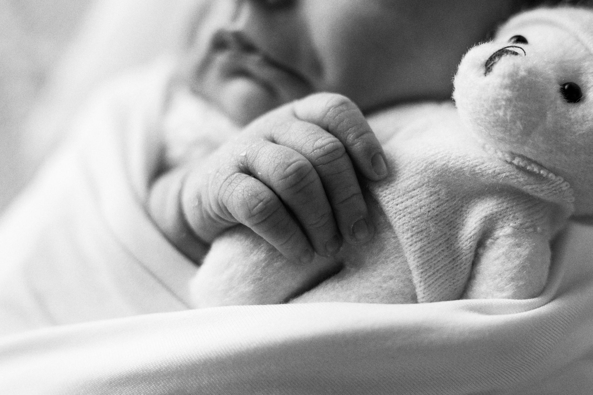 Manina chiusa a pugno posata su un lenzuolo chiaro accanto al muso di un orsetto, in bianco e nero
