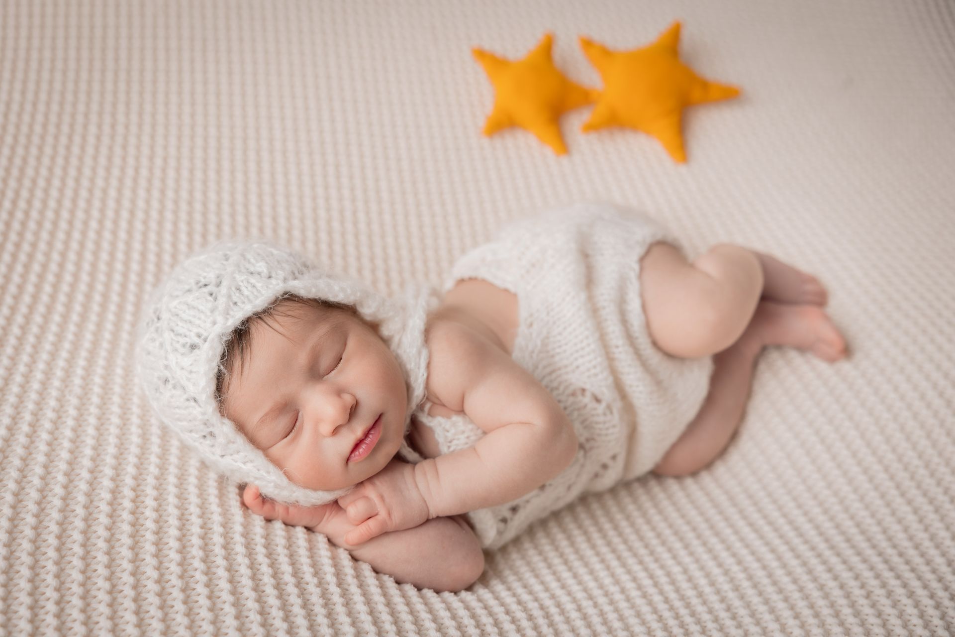 Neonato con la cuffietta di lana bianca dorme raccolto accanto a due stelline color senape, su una coperta panna a coste