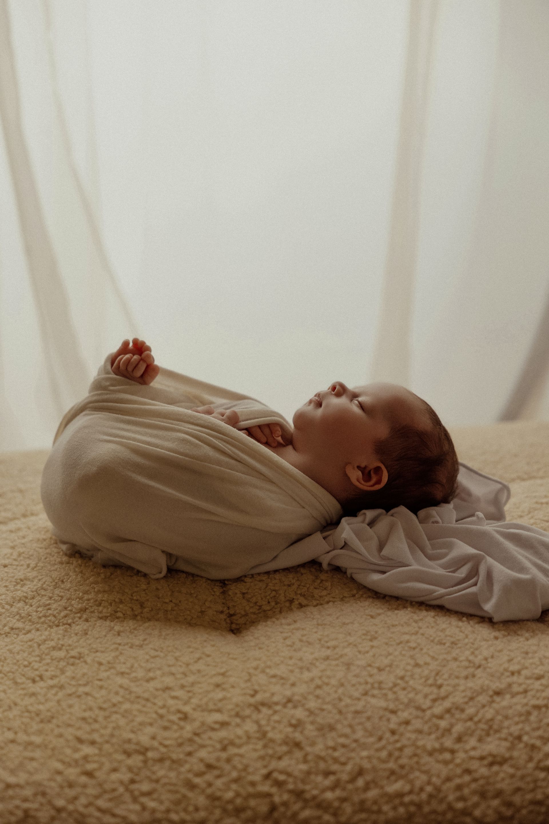 Neonato fasciato in beige riposa su un tappeto bouclé con tende bianche luminose alle spalle