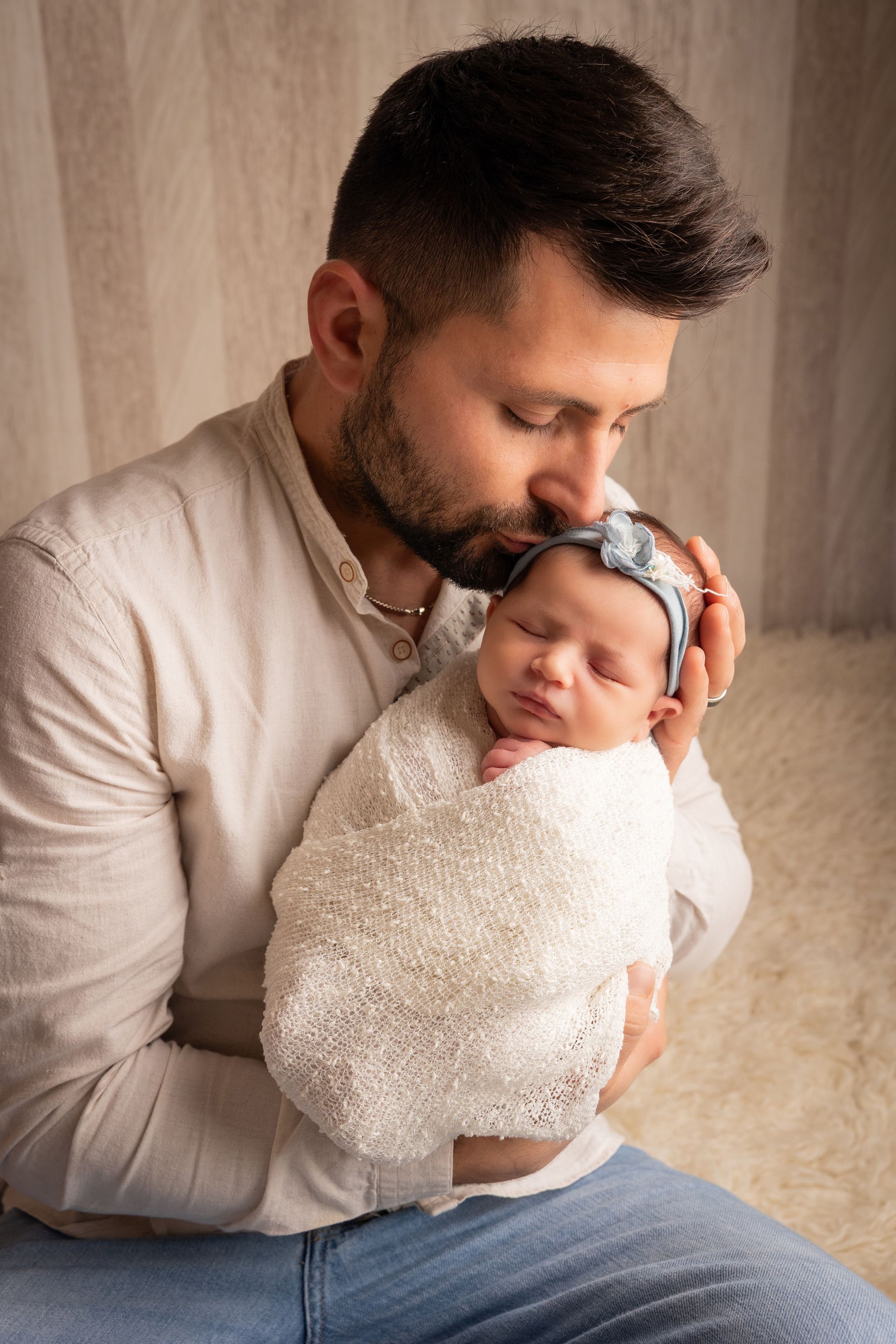 Papà in camicia beige stringe al petto la neonata fasciata e le bacia dolcemente la fronte