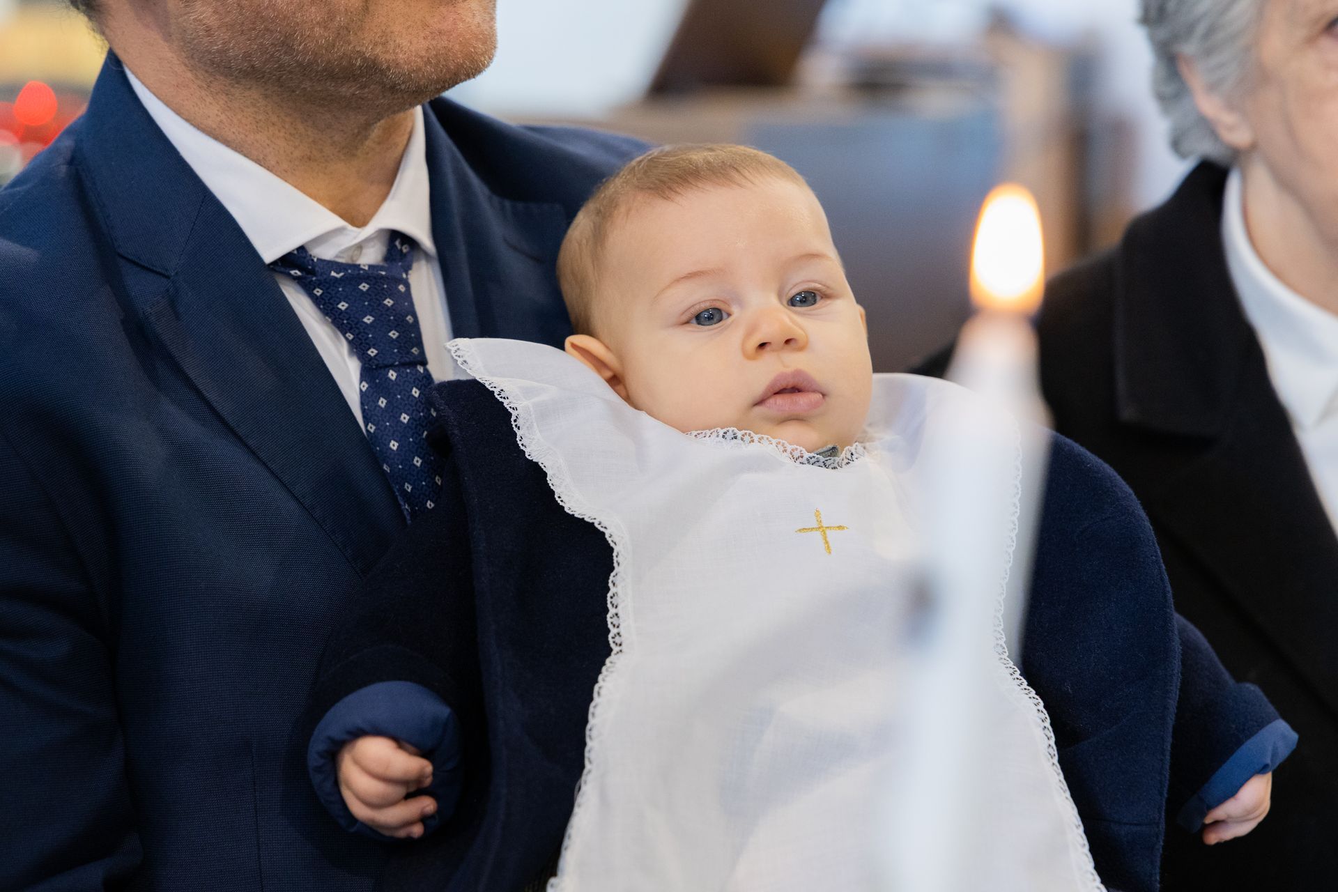 Il papà in giacca blu tiene in braccio la neonata in abito da battesimo con la croce ricamata, accanto alla candela accesa in chiesa